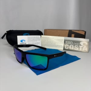 May include: Black sunglasses with blue and green mirrored lenses rest on a blue microfiber cloth. A black case, a cardboard box, and informational cards are also present. The sunglasses have the brand name "COSTA".