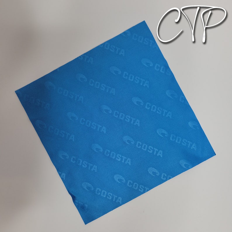 Op de afbeelding: Een vierkante, helderblauwe doek met het woord "COSTA" herhaald in een patroon over het oppervlak. De doek heeft een licht getextureerd uiterlijk en is geplaatst tegen een neutrale achtergrond. De witte letters "CYP" staan in de rechterbovenhoek.