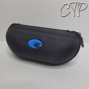May include: Black, oval-shaped sunglasses case with a textured exterior and a blue logo. The case has a zipper closure and is designed to protect eyewear. The logo is a blue wave design.