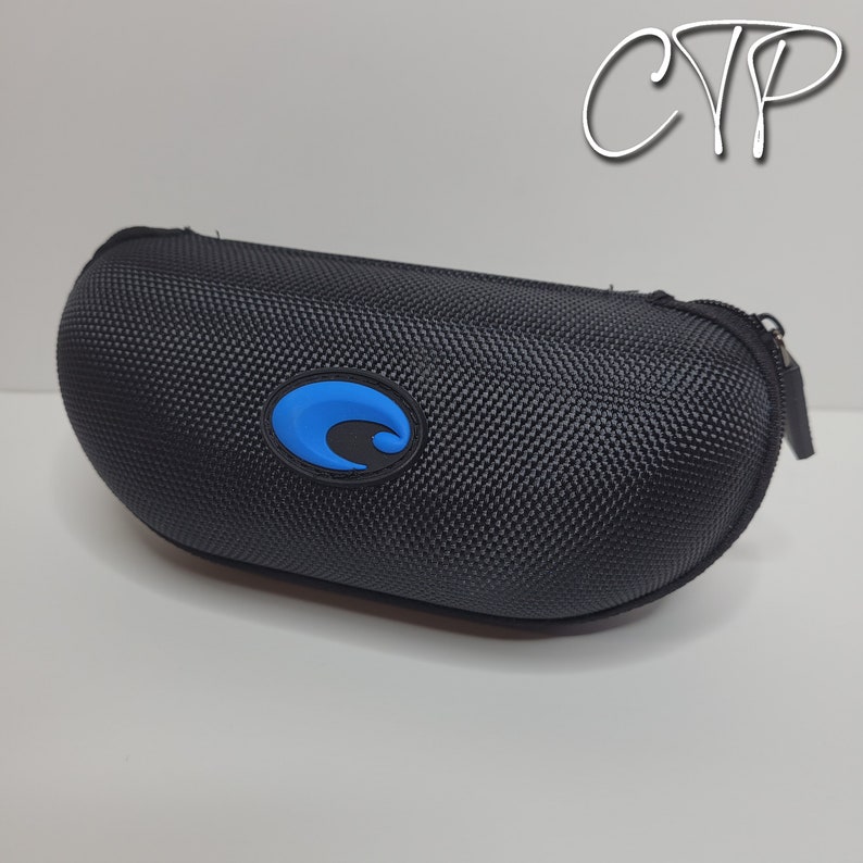 May include: Black, oval-shaped sunglasses case with a textured exterior and a blue logo. The case has a zipper closure and is designed to protect eyewear. The logo is a blue wave design.