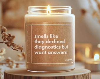 Può includere: Una candela di cera di soia con un'etichetta marrone che recita "smells like they declined diagnostics but want answers" e "crudewids". La candela è accesa e la fiamma è visibile. La candela è su una superficie di legno.
