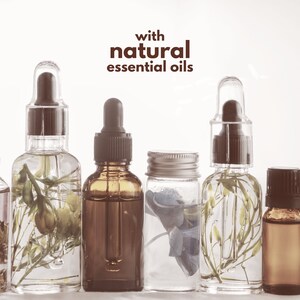 Op de afbeelding: Een rij glazen flessen met pipetten, met heldere vloeistoffen en botanische elementen. De achtergrond is wit. De tekst "with natural essential oils" wordt in bruin weergegeven. De flessen vari&euml;ren in grootte en vorm.