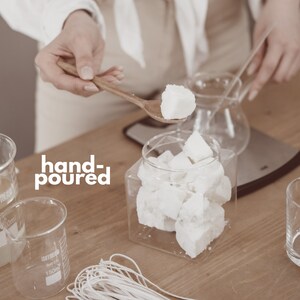 Op de afbeelding: Een persoon houdt een houten lepel vast met een wit wasblokje boven een glazen pot gevuld met witte wasblokjes. De afbeelding toont ook bekers, lonten en de tekst "hand-poured". De sc&egrave;ne suggereert kaarsen maken.