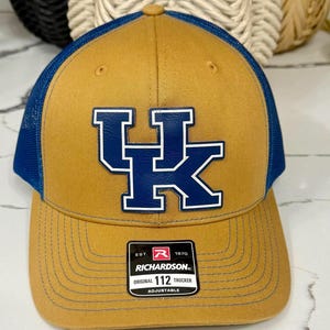 May include: A tan and blue trucker hat with the letters "UK" in blue and white on the front. The hat has a blue mesh back and a curved brim. A small black and white tag reads "RICHARDSON ORIGINAL 112 TRUCKER ADJUSTABLE".