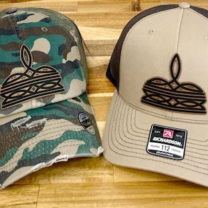 May include: Two trucker hats with a brown and black logo of a stylized burger. One hat is camouflage and the other is tan with a black mesh back. The tan hat has a Richardson 112 label.