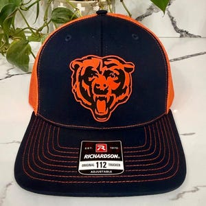 May include: Navy blue and orange trucker hat featuring a bear logo. The hat has a mesh back and a curved brim. The front panel displays an orange bear head with a black outline. The hat includes a Richardson 112 Trucker tag.