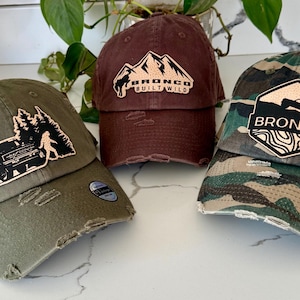 May include: Three distressed baseball caps in olive green, maroon, and camouflage patterns. Each cap features a unique patch with outdoor-themed designs and the word "BRONCO". The caps have a vintage, worn look.