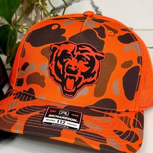 May include: An orange and brown camouflage trucker hat with a black bear head patch. The hat features a mesh back and an adjustable strap. The Richardson brand label is visible on the front.