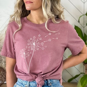 May include: A heather mauve t-shirt with a white graphic of two dandelions made of dice. The shirt is tied at the waist and the model is wearing blue denim shorts.