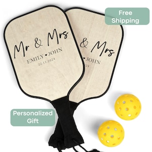 May include: Two wooden paddleball rackets with black trim and black handles. The rackets are personalized with the text "Mr. & Mrs. Emily ♥ John 22.11.2024" and "& Mrs. John 22". Two yellow paddleballs are included.