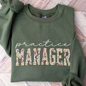 May include: A green sweatshirt with the text "practice manager" printed on the front. The text is surrounded by a floral pattern in shades of pink, white, and green.