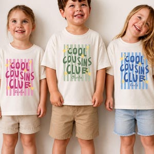 May include: Three off-white short-sleeved t-shirts featuring a vertical striped rectangular graphic with the text "COOL COUSINS CLUB" and small yellow star accents. Each shirt displays a unique colour scheme: pink, green, or blue stripes.