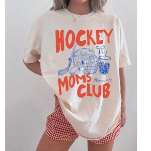 May include: Cream-colored t-shirt with the words "HOCKEY MOMS CLUB" in orange, along with blue hockey-themed graphics. The shirt also features the name "Olivia #10". The person is wearing red and white checkered shorts.