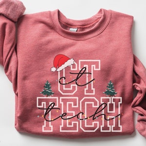 Può includere: Una felpa rosso scarlatto con la scritta "CT TECH" in lettere bianche, un cappello di Babbo Natale, due alberi di Natale e luci a stringa. Le maniche sono arrotolate. La felpa è un capo festivo.