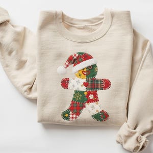 May include: A sand-colored sweatshirt featuring a patchwork gingerbread man design. The gingerbread man wears a Santa hat and is made of red, green, and white patterned fabric. The sweatshirt has long sleeves.