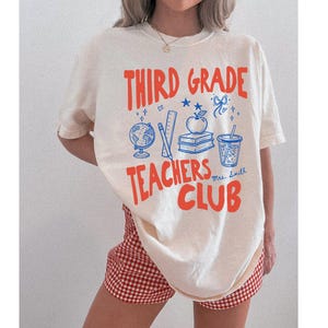 May include: Cream-colored t-shirt with a red and blue graphic that reads "Third Grade Teachers Club." The design includes a globe, ruler, apple, books, and a drink. The shirt is paired with red and white checkered shorts.