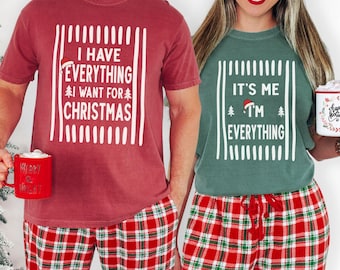 May include: A crimson t-shirt with white text reading I HAVE EVERYTHING I WANT FOR CHRISTMAS and a light green t-shirt with white text reading IT'S ME I'M EVERYTHING, both featuring small Christmas tree and Santa hat graphics, paired with red and green plaid pajama pants.