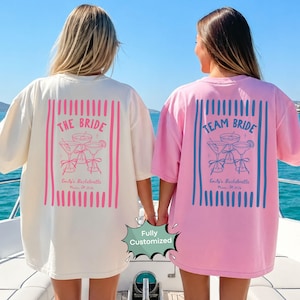 Op de afbeelding: Een off-white t-shirt met roze verticale strepen en een martini-glasafbeelding met de tekst THE BRIDE, en een roze t-shirt met blauwe verticale strepen en een martini-glasafbeelding met de tekst TEAM BRIDE, beide met aangepaste tekst.