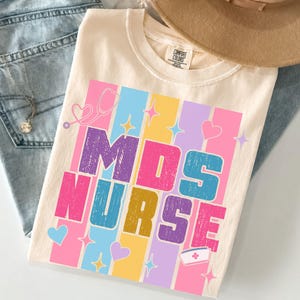 May include: Cream-colored t-shirt with the text "MDS NURSE" in colorful block letters. The design includes hearts, stars, a stethoscope, and a nurse's cap. The shirt is paired with blue jeans and a tan hat.