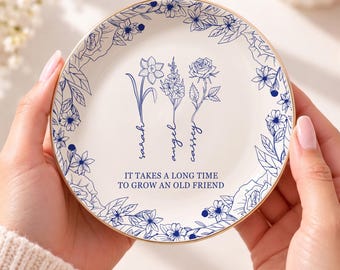 May include: A round white ceramic trinket dish with a gold-toned rim, featuring a blue floral border and three illustrated flowers with the names Sarah, Angel, and Cassey, plus the text IT TAKES A LONG TIME TO GROW AN OLD FRIEND.