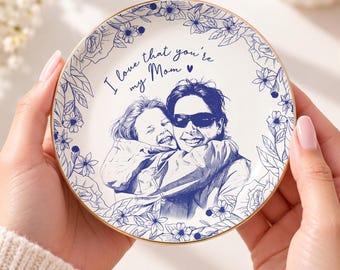 May include: A round white ceramic trinket dish featuring a blue ink illustration of a person hugging another person. The text "I love that you're my Mom" is printed in blue script, surrounded by a detailed blue floral border.