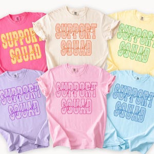 Può includere: Una collezione di sei magliette a maniche corte con girocollo in vari colori pastello, tra cui rosa, crema, giallo, lavanda, rosa chiaro e azzurro. Ogni maglietta presenta la scritta SUPPORT SQUAD stampata con un carattere in grassetto.