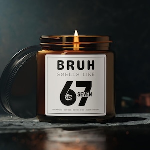 May include: A lit candle in a brown glass jar with a black lid. The label reads "BRUH SMELLS LIKE 67" in a bold font. The candle is on a dark surface with a blurred background.