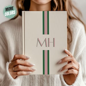 May include: A cream-colored notebook with vertical green and pink stripes and the initials "MH". The notebook is held by a person wearing a white sweater. A circular sticker in the upper left corner reads "MORE COLORS".