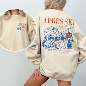 May include: Cream-coloured sweatshirt with a graphic on the front and back. The back displays a mountain scene with ski lifts, a cabin, and the text "APRÈS SKI Club BRECKENRIDGE JAN 2026". The front has a smaller version of the design.