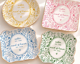 May include: Four ceramic trinket dishes with gold-toned rims, each featuring a central oval frame with the text 'sister EST. 1998 maid of honor EST. 2026' and a small heart. The dishes have yellow, pink, blue, and green botanical patterns.