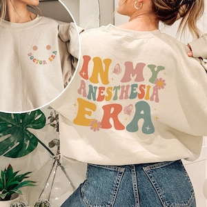 May include: A light beige sweatshirt with the text "In My Anesthesia Era" in colourful retro lettering with flowers and stars. The back of the sweatshirt has the text "Doctor Era" in a smaller font.
