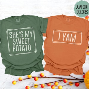 May include: Two Comfort Colors t-shirts, one green and one orange. The green shirt has the text "SHE'S MY SWEET POTATO" inside a white rectangle. The orange shirt says "I YAM" in a white rectangle. The shirts are on a white wood background.