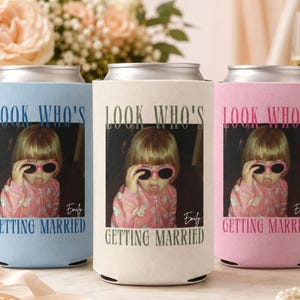 Op de afbeelding: Drie cilindrische blikkoelers van schuim in lichtblauw, crème en roze. Elk exemplaar bevat een foto van een kind met een roze zonnebril en een roze jas, met de tekst "LOOK WHO'S GETTING MARRIED" en de naam "Emily" onder de foto.