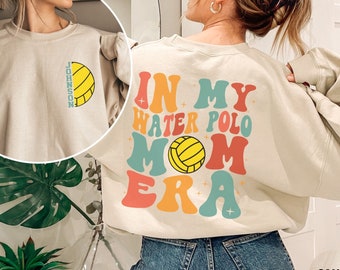 May include: A light beige sweatshirt with the text "In My Water Polo Mom Era" in colorful retro font. A yellow water polo ball is featured in the text. The sweatshirt has a relaxed fit and long sleeves.