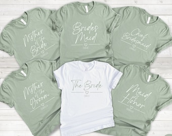 Puede incluir: Seis camisetas de color verde salvia con texto blanco. Las camisetas dicen "Madre de la novia", "Dama de honor", "Dama de honor principal", "Madre del novio", "La novia" y "Dama de honor".
