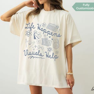 Peut inclure: T-shirt ivoire avec l'inscription bleue "Life Happens Visuals Help" et des illustrations de gribouillis. Le t-shirt a une coupe ample et des manches courtes. Les mots "Fully Customizable" sont également visibles.