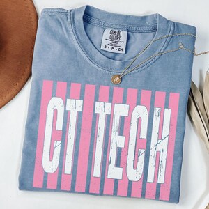 Könnte beinhalten: Ein blaues Kurzarm-T-Shirt mit einem abgenutzten weißen Aufdruck, der CT TECH über einem Hintergrund aus vertikalen rosa Streifen liest. Das T-Shirt hat ein Comfort Colors Markenetikett am inneren Halsausschnitt und ist in Größe S.