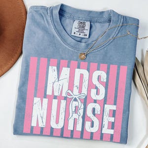 Può includere: Una t-shirt blu a maniche corte con una grafica a strisce verticali rosa e il testo bianco dall'aspetto usurato MDS NURSE con una piccola icona a forma di fiocco bianco al centro. La maglietta presenta un'etichetta Comfort Colors sul collo.