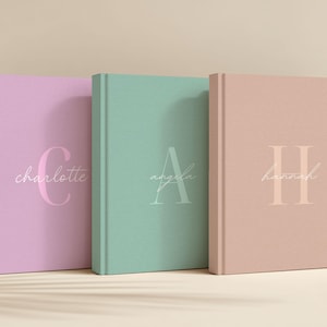 May include: Three hardcover journals in pastel colors: lavender, mint green, and peach. Each journal features a large initial and a name in a delicate script. The journals are standing upright, with the spines facing the viewer.