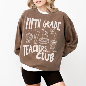 May include: Brown Fifth Grade Teachers Club sweatshirt with white graphic text and illustrations of school supplies. The design includes a globe, pencil, ruler, apple, books, and a drink. The sweatshirt is worn with black shorts.