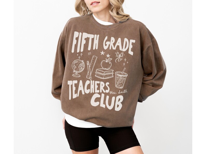 May include: Brown Fifth Grade Teachers Club sweatshirt with white graphic text and illustrations of school supplies. The design includes a globe, pencil, ruler, apple, books, and a drink. The sweatshirt is worn with black shorts.