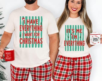 May include: Two off-white t-shirts featuring green vertical stripes and red text. The left shirt reads I HAVE EVERYTHING I WANT FOR CHRISTMAS. The right shirt reads IT'S ME I'M EVERYTHING. Both shirts are paired with red and green plaid pajama pants.
