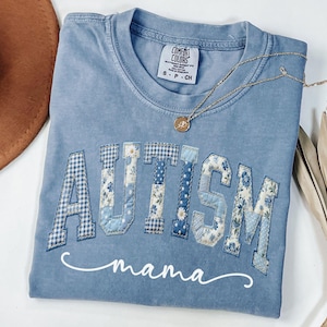 May include: A light blue t-shirt with the word "AUTISM" in patchwork letters, featuring various blue patterns. Below the word is the cursive text "mama". A gold necklace and a brown hat are also visible.