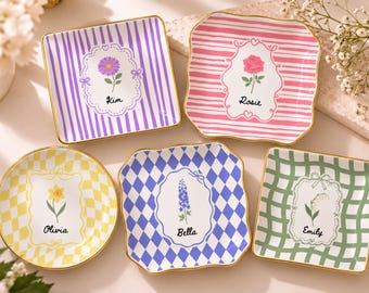 May include: Five small ceramic trinket dishes with gold-painted rims. Each dish features a unique pattern: purple stripes, pink stripes, yellow checkers, blue diamonds, and green plaid. Each center displays a floral illustration and a name.
