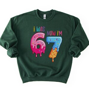 May include: Dark green sweatshirt with the text "I WAS 6, NOW I'M 7" in colorful, playful lettering. The numbers are designed to look like donuts and ice cream cones, with pink, blue, and yellow frosting.