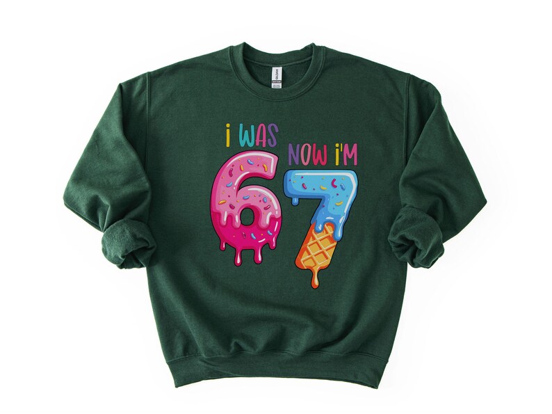 May include: Dark green sweatshirt with the text "I WAS 6, NOW I'M 7" in colorful, playful lettering. The numbers are designed to look like donuts and ice cream cones, with pink, blue, and yellow frosting.