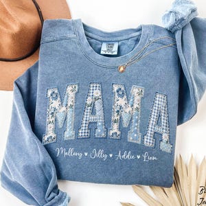 May include: Blue jean-colored sweatshirt with the word "MAMA" in appliqued letters. The letters are a mix of floral and gingham patterns. Below the word "MAMA" are the names Mallory, Jilly, Addie, and Liam. A gold necklace is draped on the sweatshirt.