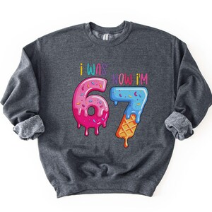 May include: Dark gray sweatshirt with the text "I WAS NOW I'M 67". The numbers are designed to look like ice cream and a donut. The number 6 is pink with sprinkles, and the number 7 is blue with sprinkles and a waffle cone.