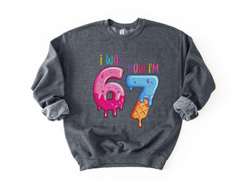 May include: Dark gray sweatshirt with the text "I WAS NOW I'M 67". The numbers are designed to look like ice cream and a donut. The number 6 is pink with sprinkles, and the number 7 is blue with sprinkles and a waffle cone.