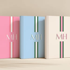 May include: Three hardcover journals in pink, light blue, and cream. Each journal features a vertical stripe design with contrasting colors and the initials "MH" embossed on the cover. The journals are displayed on a light-colored surface.